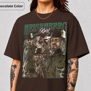 May include: Dark chocolate brown t-shirt with a graphic of a man and the text "HEISENBERG Karl" in green and white. The image shows the man in multiple poses, wearing a hat and jacket. The shirt has a crew neck.