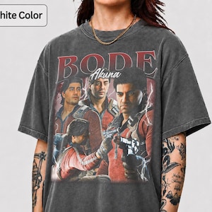 May include: A graphite-colored short-sleeve t-shirt featuring a graphic print of three figures in red jackets with the text BODE and Akuna above them. The design includes lightning effects and a distressed, washed-out fabric appearance.