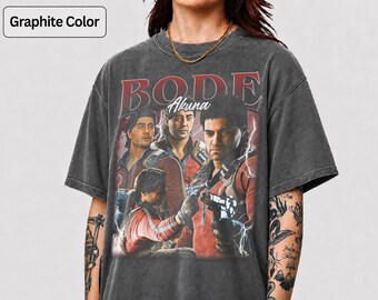 May include: A graphite-colored short-sleeve t-shirt featuring a graphic print of three figures in red jackets with the text BODE and Akuna above them. The design includes lightning effects and a distressed, washed-out fabric appearance.