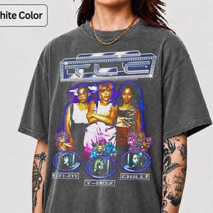 May include: A graphite-coloured short-sleeve t-shirt featuring a graphic print of the music group TLC. The design includes the text TLC at the top, images of the three members, and individual name labels for Left-Eye, T-Boz, and Chilli below.