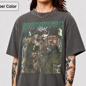 May include: Dark grey t-shirt with a graphic of a group of men and the text "HEISENBERG Karl" in green. The shirt has a vintage appearance. The model is wearing a gold chain. The t-shirt is pepper color.