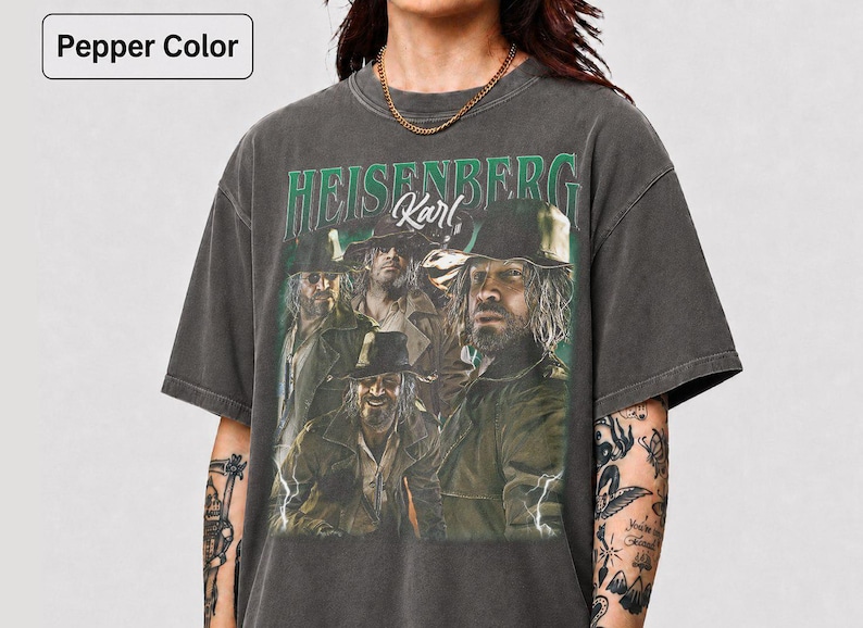 May include: Dark grey t-shirt with a graphic of a group of men and the text "HEISENBERG Karl" in green. The shirt has a vintage appearance. The model is wearing a gold chain. The t-shirt is pepper color.