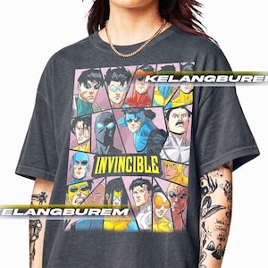 May include: Dark gray t-shirt featuring a comic book style graphic with multiple superhero characters. The word "INVINCIBLE" is printed in white. The shirt has a crew neck and short sleeves.
