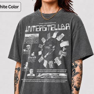 Peut inclure: T-shirt graphite avec le mot "INTERSTELLAR" et un motif de station spatiale. Le t-shirt contient également du texte sur le destin et le regard vers le ciel. Le t-shirt est à manches courtes.