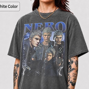 Puede incluir: Una camiseta de manga corta de color grafito con un estampado gráfico de cuatro personajes de la serie de videojuegos Devil May Cry. El texto NERO está impreso en azul sobre los personajes y la camiseta tiene un aspecto desgastado.