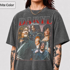 May include: A graphite-colored short-sleeve t-shirt featuring a graphic print of the character Daryl Dixon. The design includes the text DARYL DIXON in red and white lettering, with multiple images of the character holding weapons on the front.