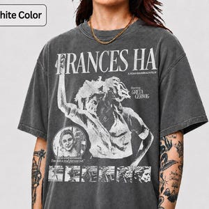 May include: Graphite-colored t-shirt featuring the movie title "FRANCES HA" in large white letters. The shirt includes a graphic of a woman and the text "A Noah Baumbach Film" and "Starring Greta Gerwig". The shirt also has the text "I'm so embarrassed. I'm not a real person yet."