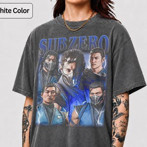Op de afbeelding: Grafietkleurig t-shirt met een afbeelding van Sub-Zero-personages. Het ontwerp bevat het woord "SUBZERO" in blauw, met figuren in blauwe en witte maskers. Het shirt heeft een vintage, versleten uitstraling.
