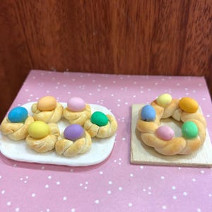 Op de afbeelding: Miniatuurgebak in paasthema. Zes kleine, ronde gebakjes met kleurrijke eivormige snoepjes op een wit bord. Een gevlochten broodkrans met kleurrijke eieren ligt op een houten plank.