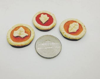 May include: Four miniature pie-shaped decorations with a leaf design, in shades of orange, red, and cream. A silver coin is next to the pies. The pies have a dark brown base and a light-colored crust.