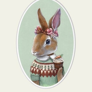 Op de afbeelding: Een ingelijste illustratie van een bruin konijn dat een groene trui draagt met een witte kraag en een bloemenkrans. Het konijn houdt een cupcake vast met een wortel erop. De achtergrond is een lichtgroene ovaal.