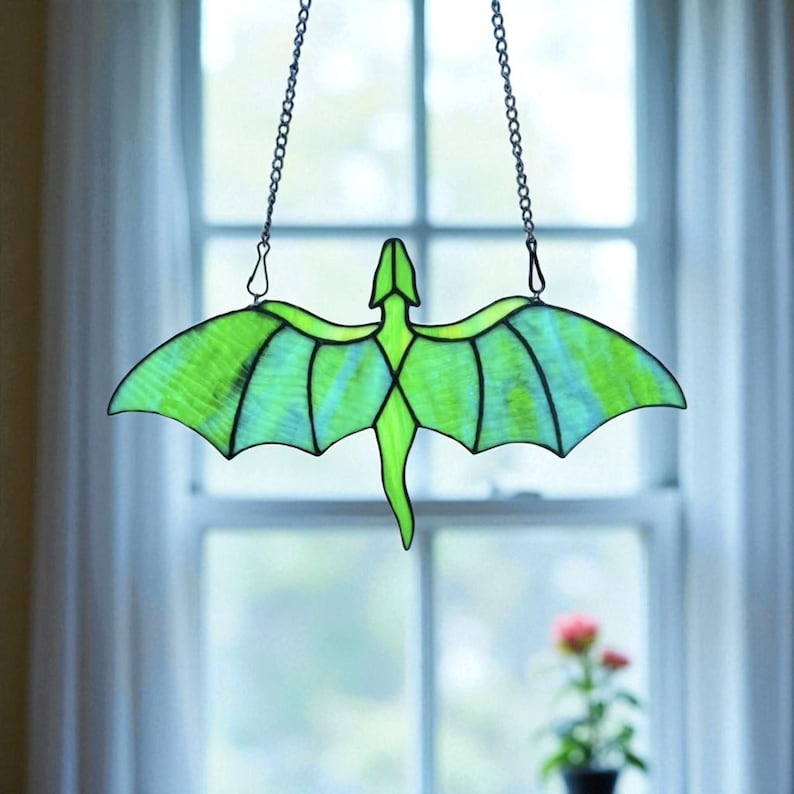 May include: A stained glass dragon with outstretched wings, in shades of green and blue, hangs from a chain. The dragon's body and wings are outlined in black, creating a striking contrast. The artwork is displayed in front of a window.