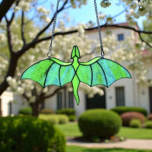 May include: A stained glass dragon suncatcher with a green and blue color scheme. The dragon is suspended by a silver chain. The wings are spread wide, and the suncatcher is hanging in front of a blurred background of trees and a house.