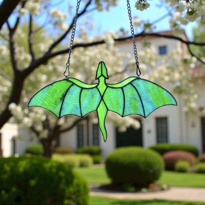 May include: A stained glass dragon suncatcher with a green and blue color scheme. The dragon is suspended by a silver chain. The wings are spread wide, and the suncatcher is hanging in front of a blurred background of trees and a house.