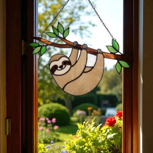 May include: Stained glass sloth hanging from a branch with green leaves. The sloth is light brown with a white belly and dark brown details. The branch is brown, and the leaves are green. The piece hangs from a chain, and is displayed in a window.