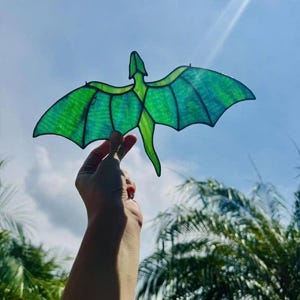 May include: A green dragon-shaped suncatcher with outstretched wings, held against a bright blue sky. The dragon has a detailed design with varying shades of green, creating a stained-glass effect. The wingspan appears to be approximately 6 inches.