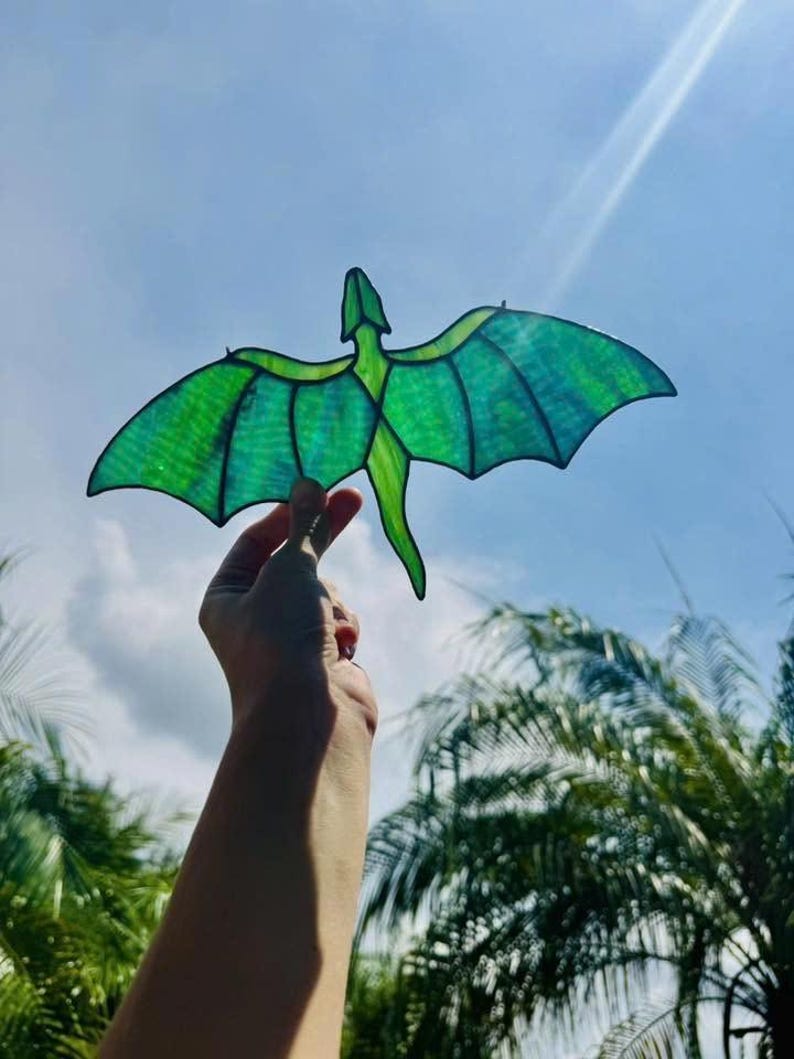 May include: A green dragon-shaped suncatcher with outstretched wings, held against a bright blue sky. The dragon has a detailed design with varying shades of green, creating a stained-glass effect. The wingspan appears to be approximately 6 inches.