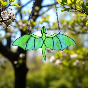 May include: A stained glass dragon suncatcher with a green body and wings, hanging from a silver chain. The dragon has a detailed design with black outlines and is suspended against a blurred background of green foliage and sunlight.