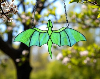 May include: A stained glass dragon suncatcher with a green body and wings, hanging from a silver chain. The dragon has a detailed design with black outlines and is suspended against a blurred background of green foliage and sunlight.