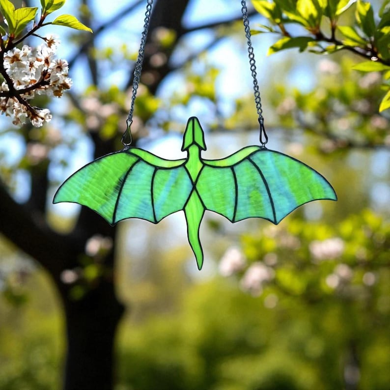 May include: A stained glass dragon suncatcher with a green body and wings, hanging from a silver chain. The dragon has a detailed design with black outlines and is suspended against a blurred background of green foliage and sunlight.