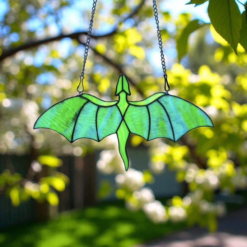 May include: A stained glass dragon suncatcher with a green body and wings, hanging from a silver chain. The dragon's wings have a striped pattern, and the suncatcher is set against a blurred background of green foliage and sunlight.