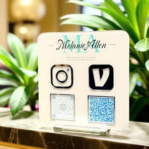 May include: A square white acrylic sign featuring the text Melanie Allen, Christian Fiction Author, and Welcome to Hope's Bluff. The sign displays a black Instagram icon, a black V icon, and two square QR codes in silver and blue.
