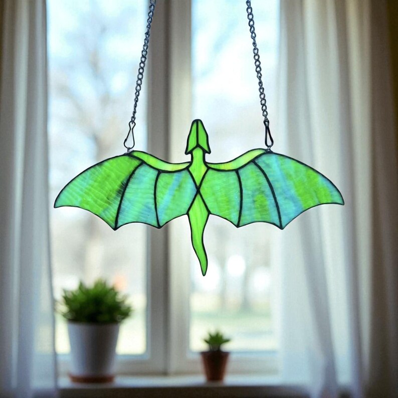 May include: A stained glass dragon suncatcher with a green and blue color scheme. The dragon is suspended by a chain and hangs in front of a window. The dragon's wings are spread, and the body is detailed with black lines.