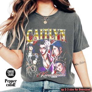 May include: Dark gray t-shirt with a graphic featuring the name "CAITLYN" in gold lettering, and illustrations of several characters. The shirt also has the text "Kiranman" and "up 2-3 size for Oversized".
