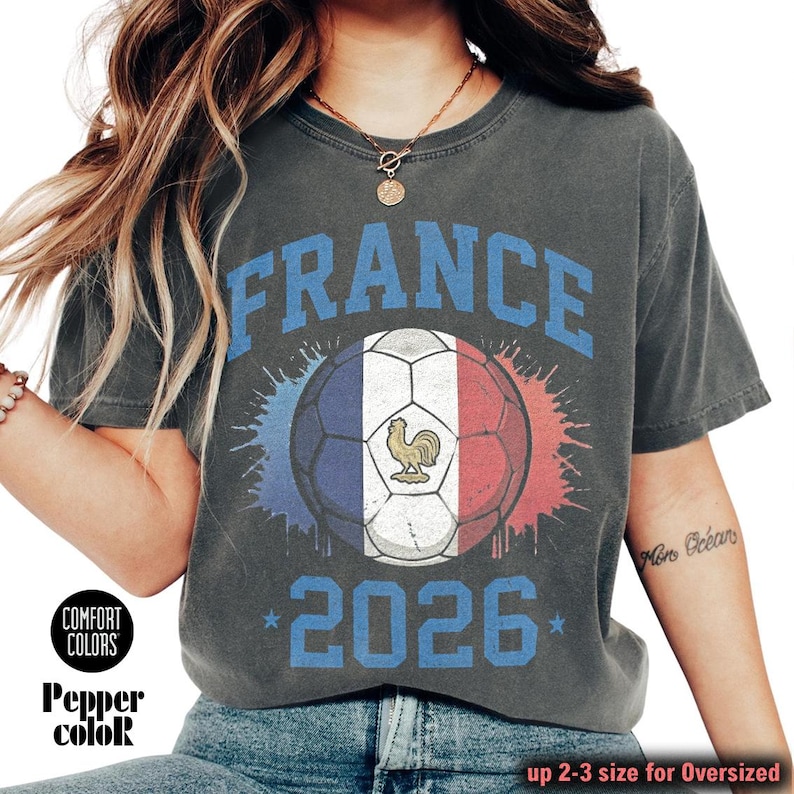 Op de afbeelding: Donkergrijs t-shirt met het woord "FRANCE" in blauw boven een voetbal in de kleuren van de Franse vlag. Het jaar "2026" staat eronder in het blauw gedrukt. Het shirt heeft ook de woorden "Comfort Colors" en "Pepper color" linksonder.