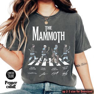 K&ouml;nnte beinhalten: Dunkelgraues T-Shirt mit dem Text "The Mammoth" &uuml;ber einer Illustration von vier Eishockeyspielern, die einen Zebrastreifen &uuml;berqueren. Die Namen der Spieler sind unter der Illustration aufgedruckt. Das Shirt tr&auml;gt auch die Aufschrift "Comfort Colors" und "Pepper color".