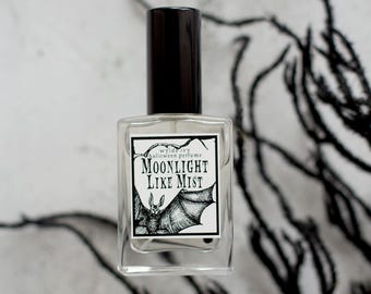 May include: A clear glass perfume bottle with a black cap. The label reads "Moonlight Like Mist" with a bat illustration. The background features black, abstract, branch-like shapes, creating a Halloween theme.