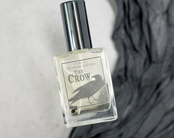 May include: A clear glass bottle of perfume with a black cap. The label reads "wyde ivy the omens collection THE CROW" and features a black crow illustration.
