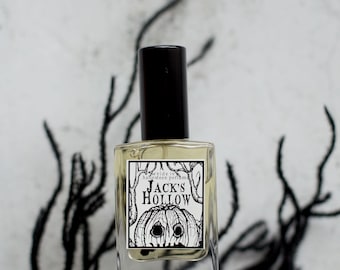 May include: A clear glass perfume bottle with a black cap. The label features a black and white illustration of a jack-o'-lantern with the text "Jack's Hollow". The background is a blurred white and gray with black decorative elements.