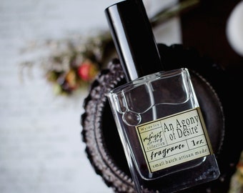 May include: A clear glass bottle of perfume with a black cap. The label is gold with black text that reads "Wyde Ivy, A Midnight Collection, An Agony of Desire, Fragrance, 1 oz, small batch artisan made".
