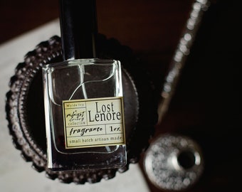 May include: A clear glass bottle with a black cap and a label that reads "Wilde Ivy Midnight Alchemy Collection Lost Lenore Fragrance 1 oz. small batch artisan made".