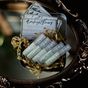 May include: A tin box filled with small glass bottles of perfume. The bottles are labeled with names like "Lost Lenore", "A Midnight Dreary", and "An Agony of Desire". The box is surrounded by gold tissue paper.