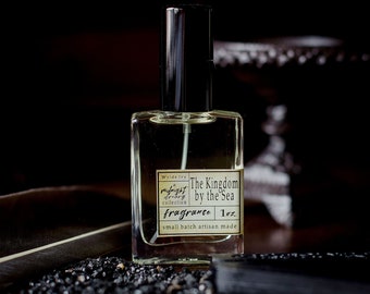 May include: A clear glass bottle of fragrance with a black cap and label. The label reads "Wyldside Ivy, Midnight Cherry Collection, The Kingdom by the Sea, Fragrance, 1 oz, small batch artisan made".