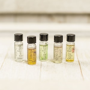 May include: Five small glass bottles with black caps and labels. The labels have different names for scents, including "Fir and Frost", "Frosted Lavender", "Rain + Fern", "Shine Like Sugar", and "Sugar Rose".