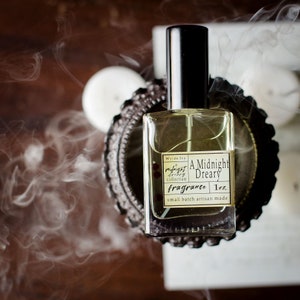 May include: A clear glass bottle of "A Midnight Dreary" fragrance from Wyde Ivy's Midnight Dreary collection. The bottle is sitting on a decorative metal stand and has a black cap. The label is white with black text and red dots. The bottle contains 1 oz of fragrance.
