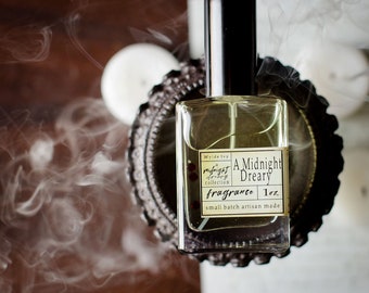 May include: A clear glass bottle of "A Midnight Dreary" fragrance from Wyde Ivy's Midnight Dreary collection. The bottle is sitting on a decorative metal stand and has a black cap. The label is white with black text and red dots. The bottle contains 1 oz of fragrance.