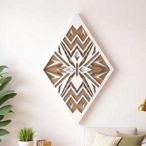 Pode incluir: Uma obra de arte de parede em forma de diamante branco com um design geométrico em camadas em tons de castanho e branco. A obra de arte é montada numa parede branca, acima de um sofá branco com almofadas verdes e creme. À esquerda, uma planta verde alta numa cesta de vime.