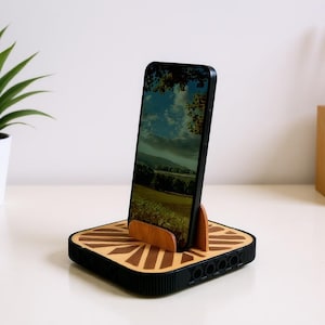 Puede incluir: Un teléfono inteligente negro se muestra en un soporte de teléfono de madera. El soporte tiene una base cuadrada con un diseño geométrico y un soporte de madera para el teléfono. A la izquierda hay una planta verde en una maceta blanca.