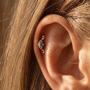 Può includere: Un orecchino a spirale color oro con una piccola gemma rotonda color verde acqua. L'orecchino ha un design delicato e a spirale. L'orecchino è posizionato sull'orecchio di una persona con capelli castano chiaro.