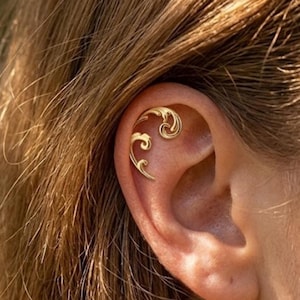 Pode incluir: Um piercing dourado com um design ornamental e giratório. A joia é colocada na cartilagem superior da orelha, apresentando uma forma curva com detalhes delicados. O piercing é realçado sobre o cabelo castanho claro.