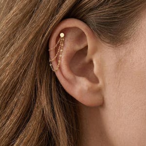 May include: A gold ear cuff with a small disc stud and three fine chain drops. The jewellery is designed for the upper ear, adding a touch of elegance. The chains hang gracefully, creating a subtle, yet eye-catching effect.