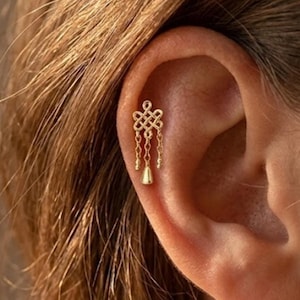 May include: A gold-toned ear piercing stud featuring an intricate, symmetrical knot design at the top. Three delicate, thin gold chains hang from the knot, with the center chain ending in a small, smooth, bell-shaped gold charm.