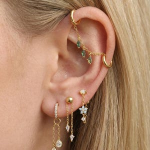 May include: Gold-toned ear jewelry set featuring multiple earrings. Includes a chain earring with green gems, a hoop earring, a stud earring with a flower, and a hoop earring with dangling teardrop-shaped gems.