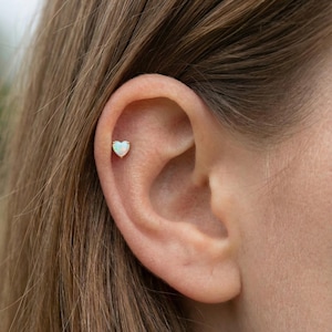 Puede incluir: Un pendiente en forma de corazón, de color dorado, con una piedra de ópalo blanco en la oreja. El pendiente es un pequeño pendiente de botón. El ópalo tiene un brillo sutil e iridiscente. La oreja está enfocada, con el pelo borroso en el fondo.