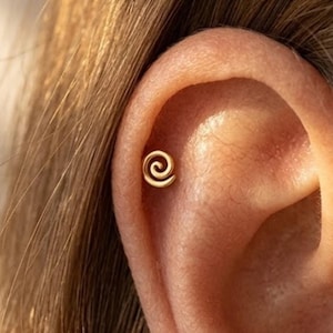 Può includere: Un orecchino a spirale color oro è mostrato nell'orecchio. L'orecchino è piccolo e ha un design minimalista. L'orecchio è di colore chiaro e sono visibili dei capelli.