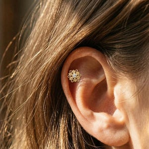 Può includere: Un orecchino a lobo dorato caratterizzato da un delicato design floreale o a mandala traforato, con una singola pietra preziosa trasparente a taglio rotondo incastonata al centro. Il gioiello è posizionato nella cartilagine superiore.